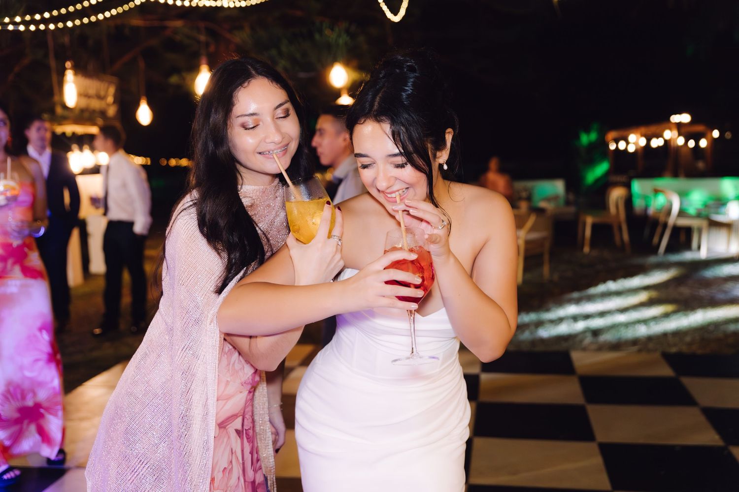 Bride and guest in pink dress clink colorful cocktails together on checkered dance floor at outdoor wedding reception.