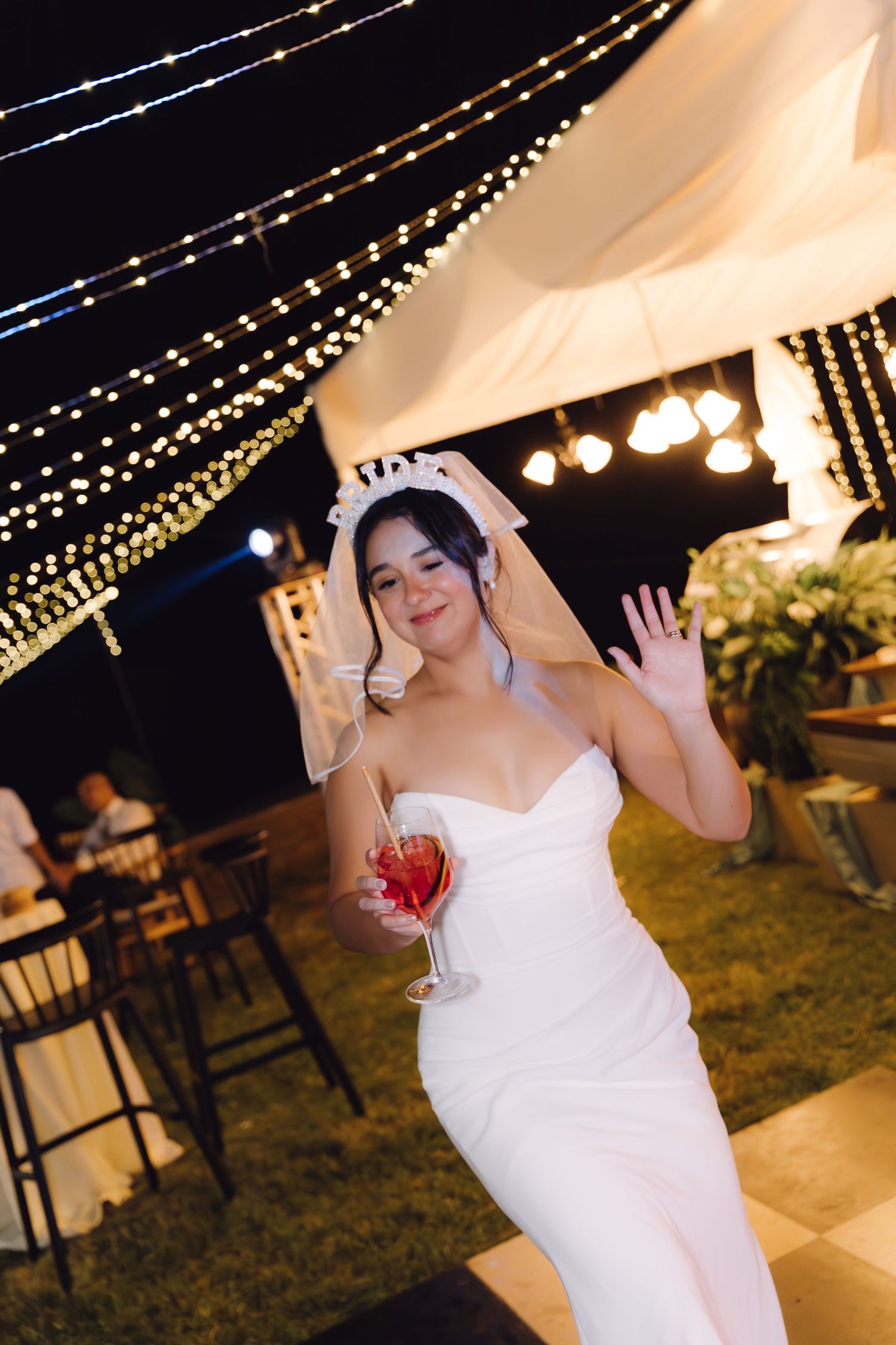 Smiling bride in white strapless gown and veil holds red wine glass on checkered dance floor under string lights.