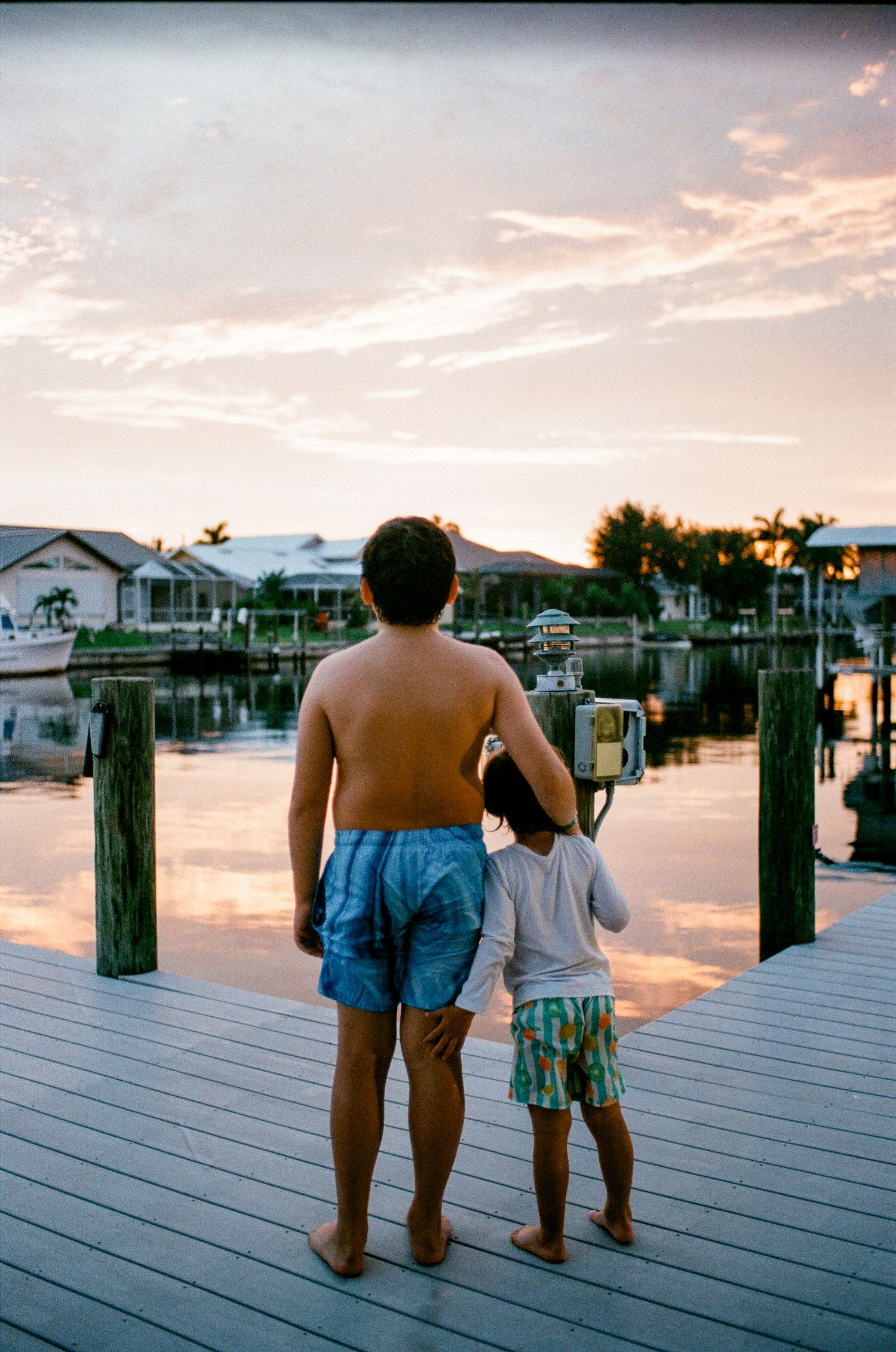 A shirtless man and young child stand on a wooden dock at sunset, overlooking a calm waterway with boats and homes.