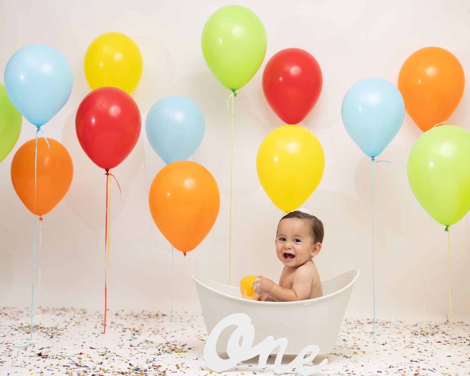 Happy baby sitting in a white tub surrounded by colorful balloons celebrating their first birthday.