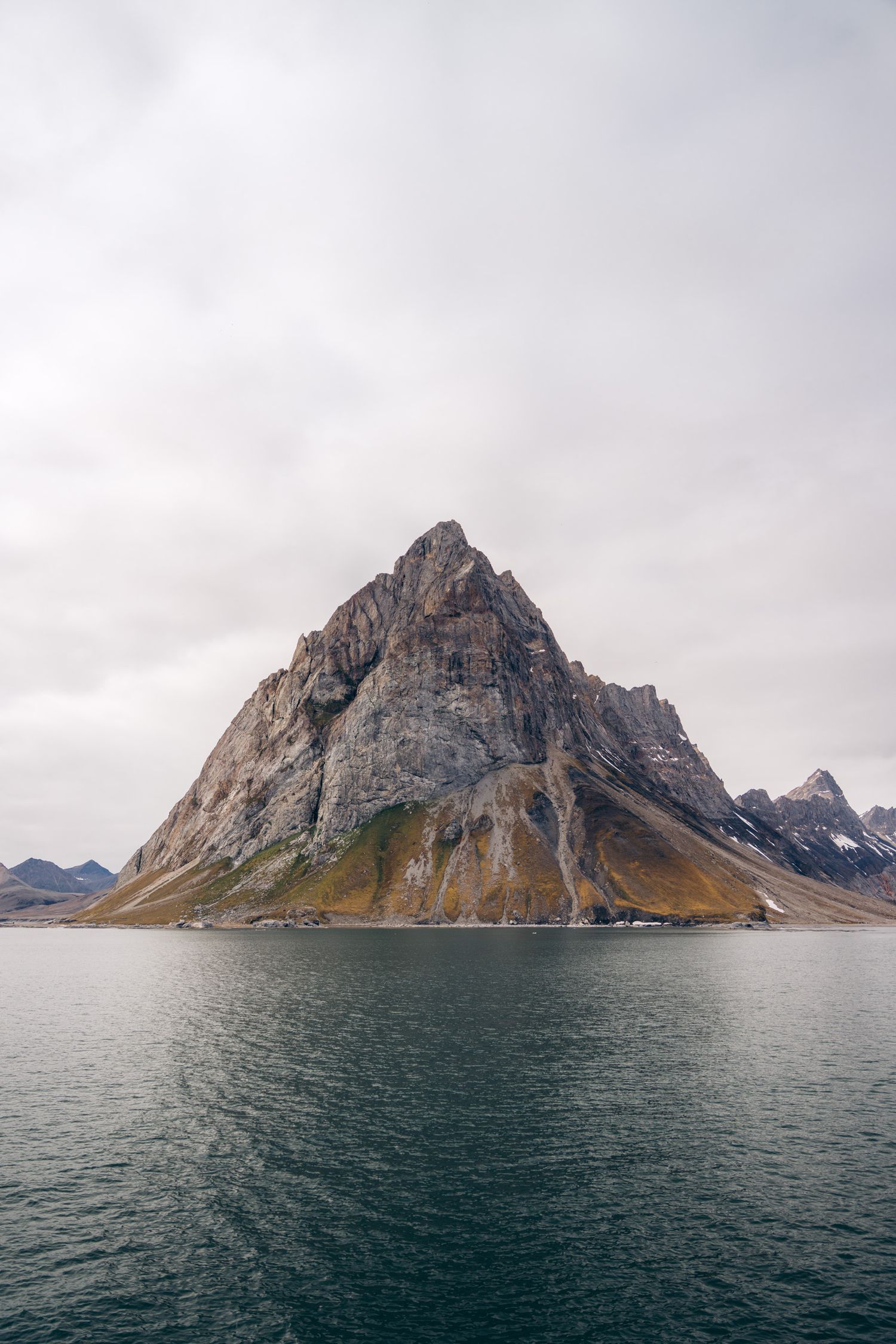 Montagne pyramidale grise et ocre plongeant dans un fjord lisse du Svalbard