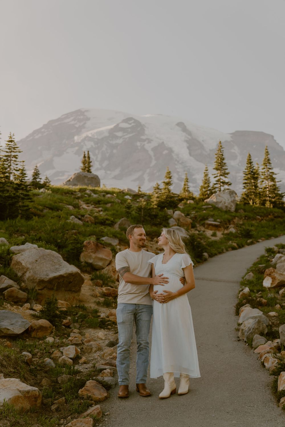 Couple holding pregnant belly on mountain path with snowy Mount Rainier backdrop.