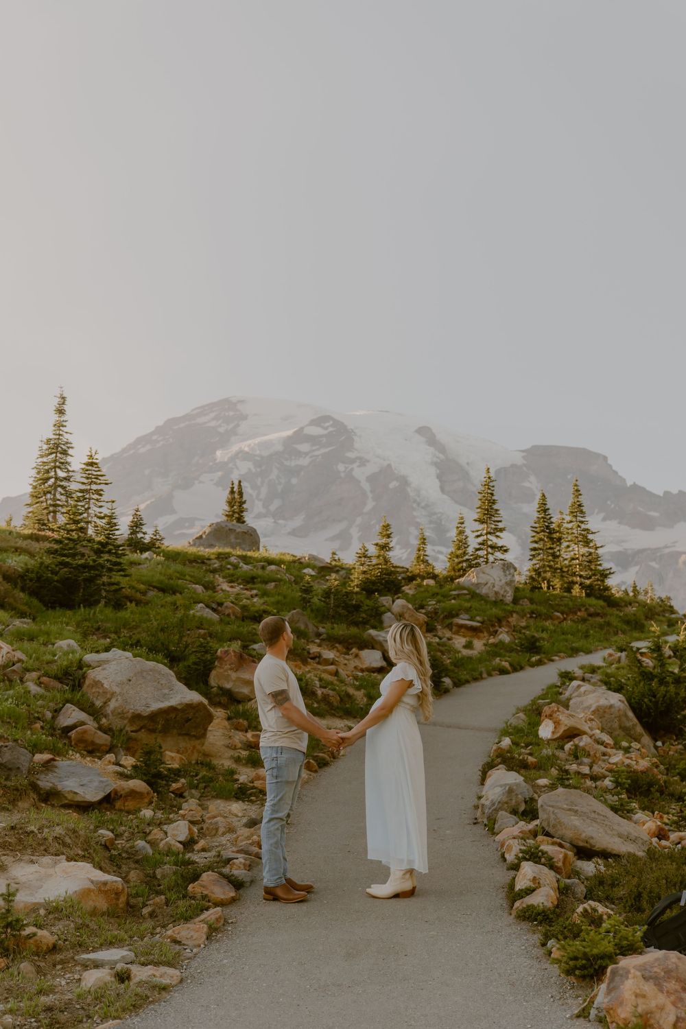 Couple holding hands on a mountain path with snow-capped peaks and evergreen trees in the background.