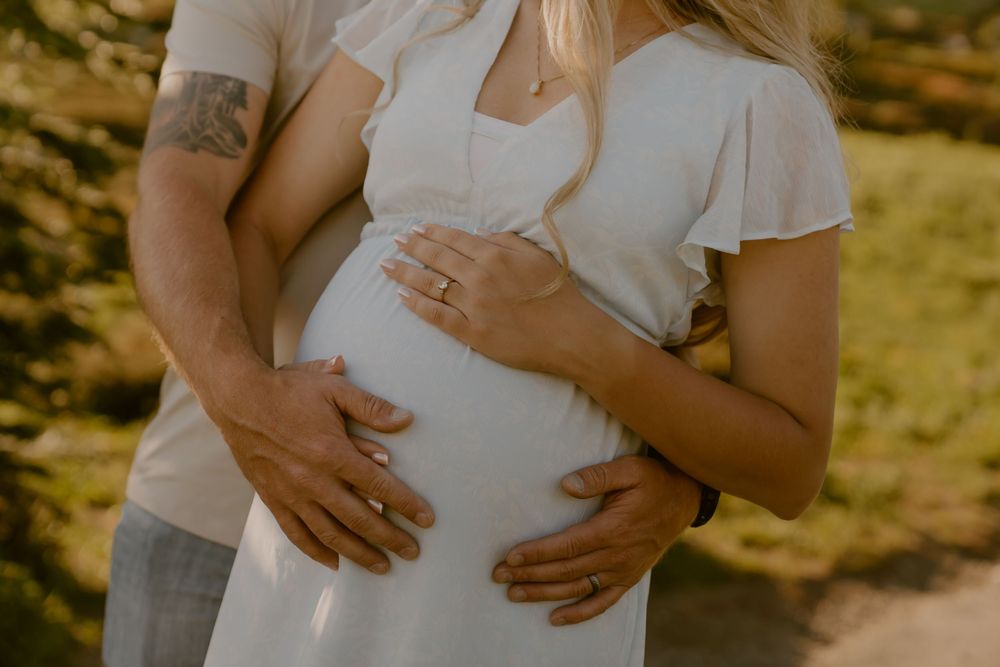 Couple cradling pregnant belly outdoors, woman in white dress with rings visible, man with tattoo embracing her.