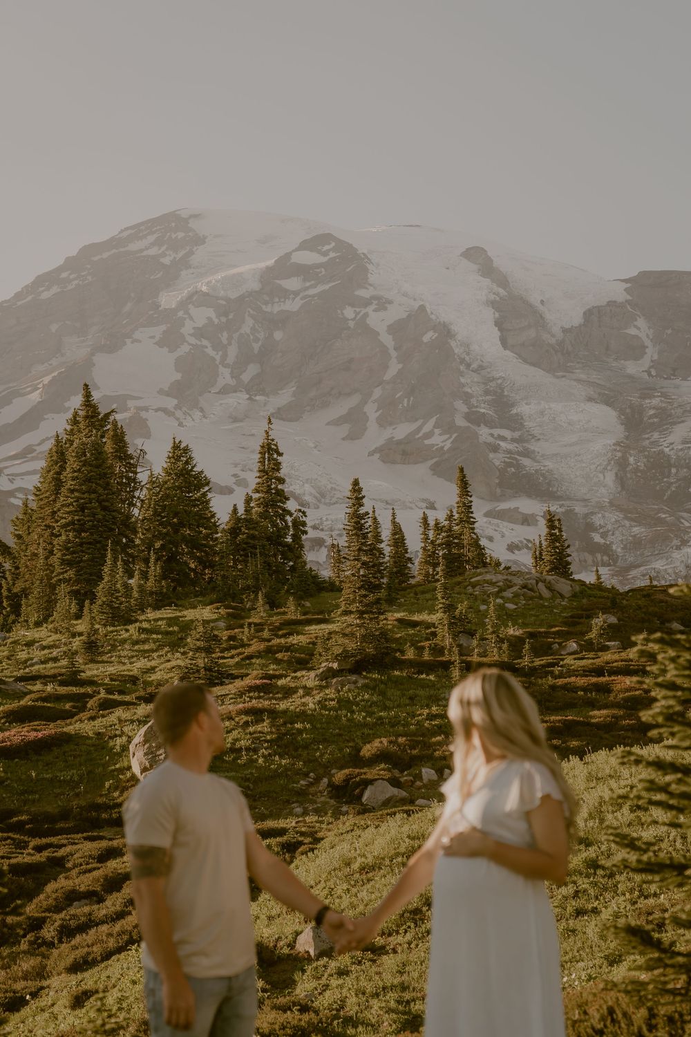 Pregnant couple holding hands on alpine meadow trail with snowy mountain peak behind.