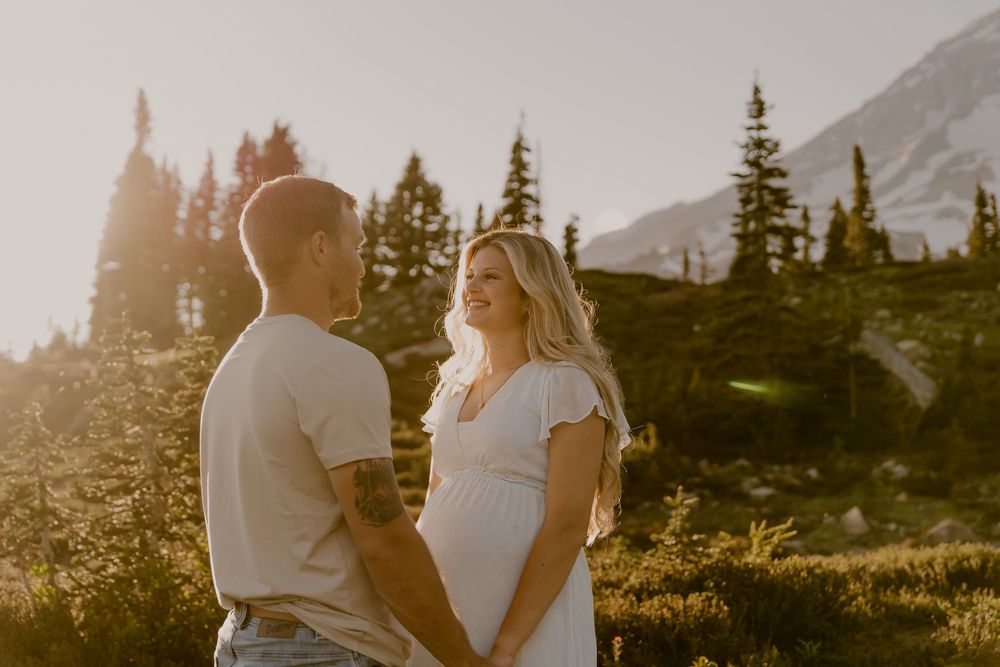 Smiling pregnant couple holding hands in golden sunset light with evergreen trees.