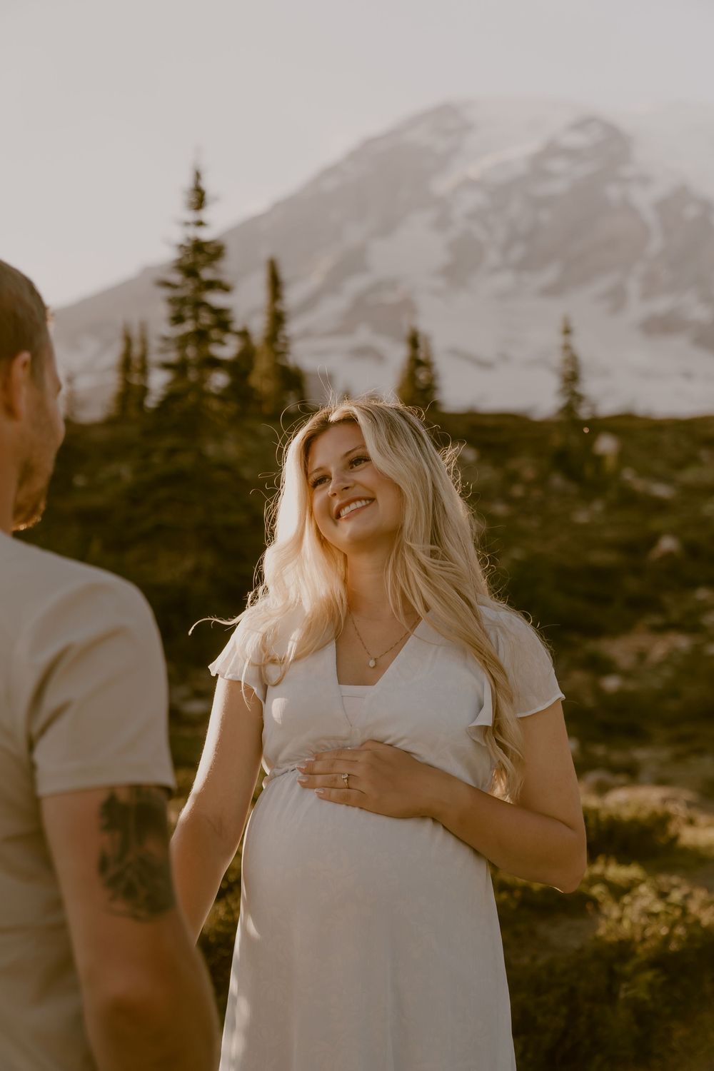 Pregnant woman in white dress smiling at partner during golden hour maternity shoot at Mount Rainier.