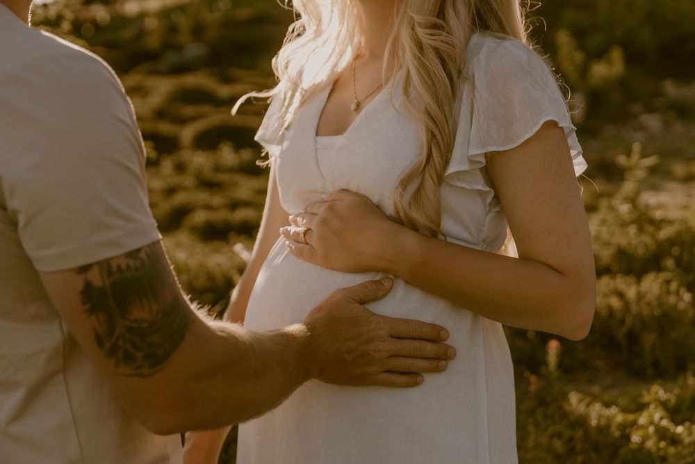 Pregnant woman in white dress with partner's tattooed hand on her baby bump outdoors in golden light.