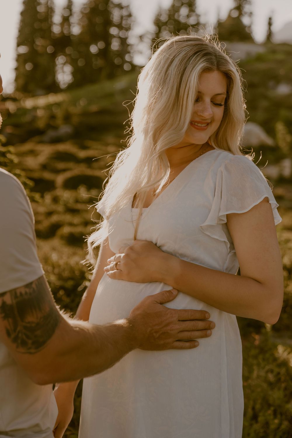 Pregnant woman in white dress cradling baby bump with partner's tattooed hand.