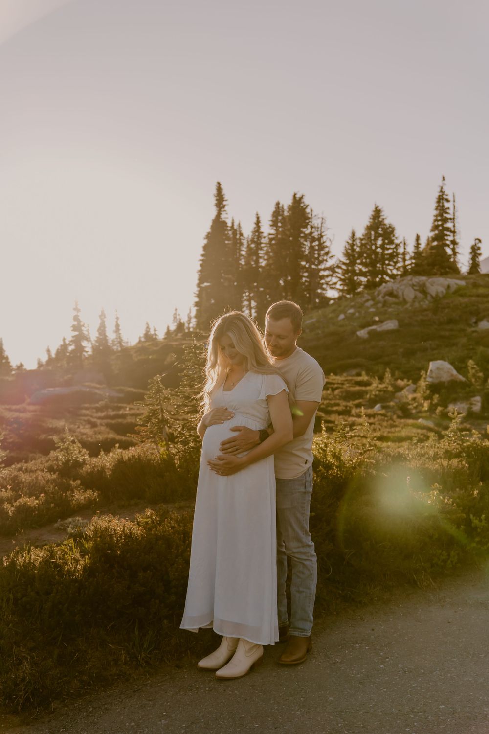 Pregnant couple embracing on mountain path at golden hour with evergreen trees backdrop.