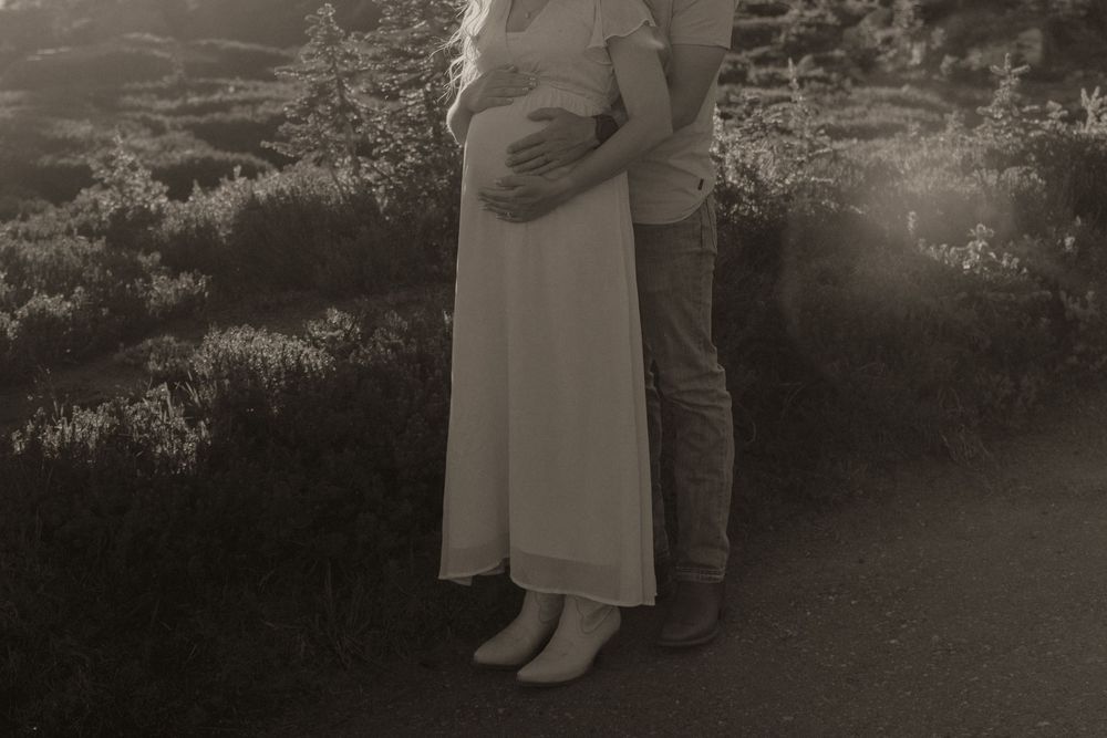Black and white photo of a pregnant couple embracing outdoors, woman in white dress cradling baby bump.