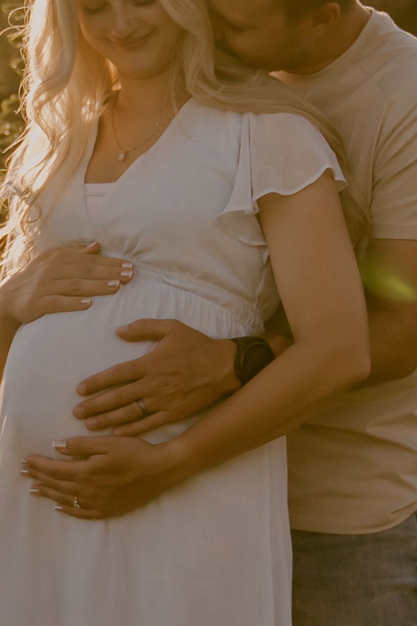 Pregnant woman in white dress with partner tenderly cradling baby bump in warm golden sunset light.