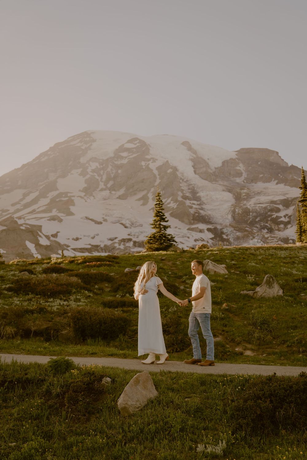 Pregnant couple holding hands on a mountain trail with snow-capped Mount Rainier in the background.
