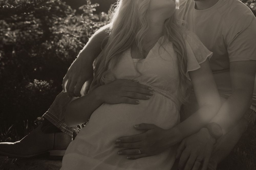 Black and white maternity photo of a couple sitting together, hands tenderly cradling a pregnant belly outdoors.