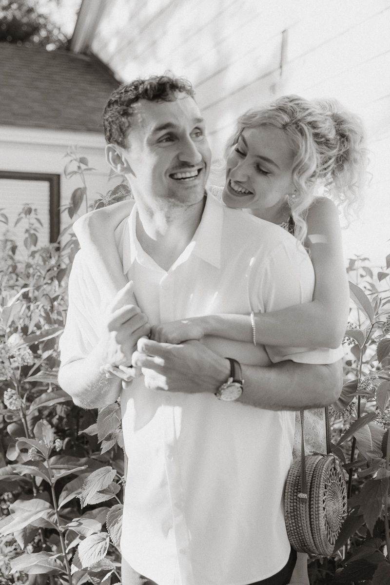 Black and white photo of a smiling couple embracing outdoors, woman hugging man from behind near lush greenery.