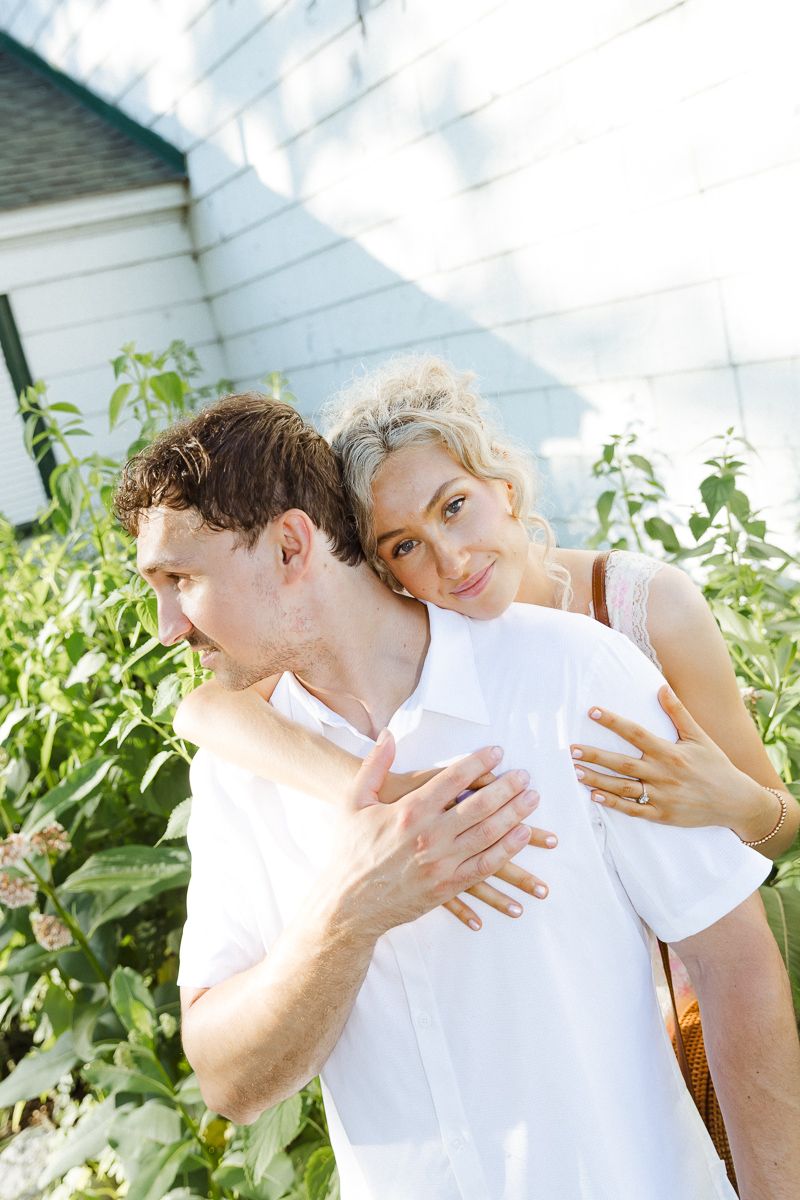 A couple embraces outdoors near green plants, woman with blonde hair rests her head on man's shoulder smiling.
