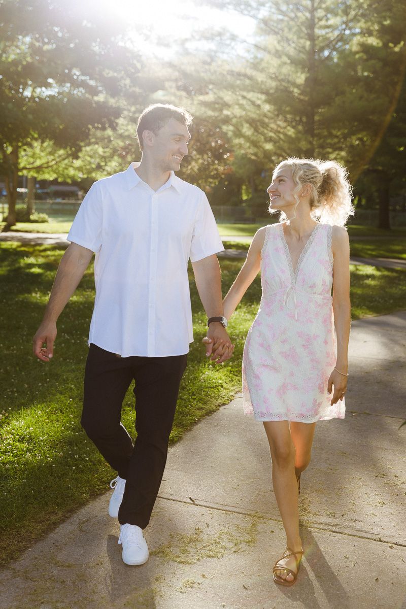 A smiling couple holds hands while walking on a sunny park path, man in white shirt and woman in floral dress.