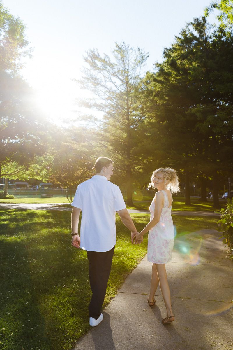 Couple holding hands walking on a sunny park path, woman in floral dress glancing back, golden sunlight through trees.