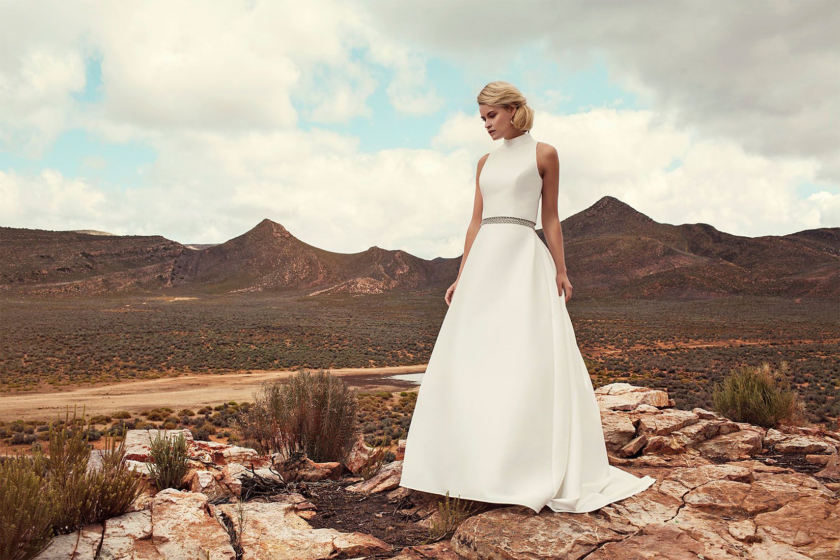 Bride wearing minimalist two-piece white wedding dress stands on rocky desert terrain with mountains behind.