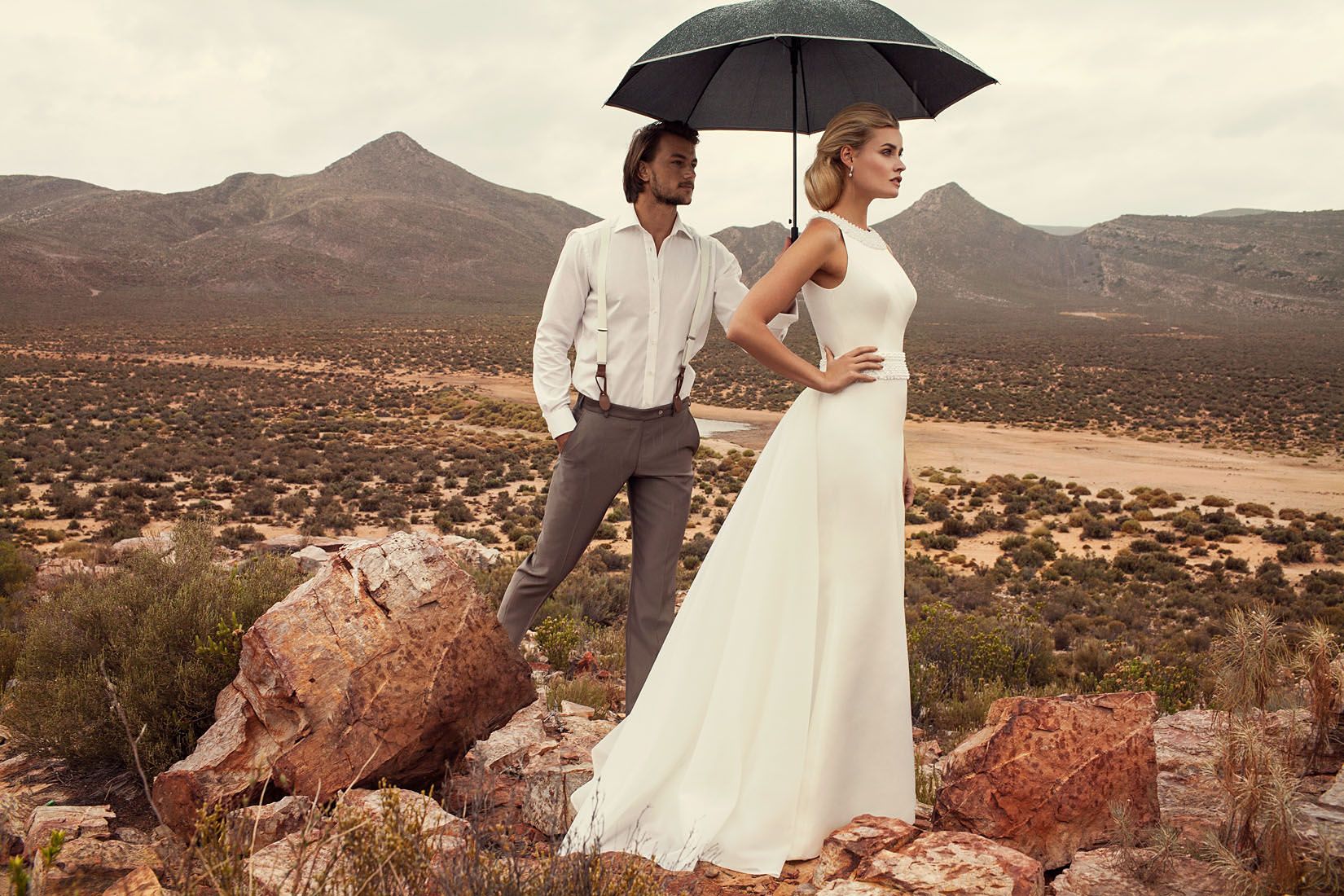 Bride and groom share umbrella in arid desert landscape, woman wearing flowing white wedding gown.