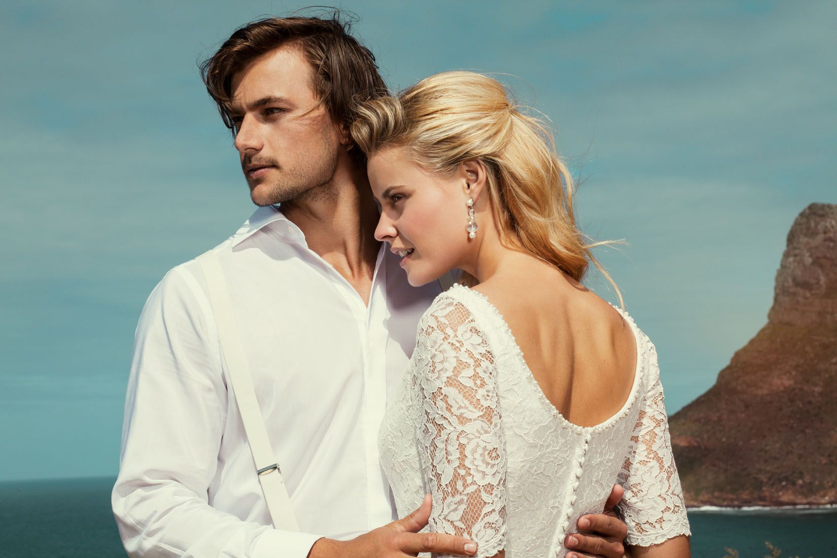 Couple in romantic embrace wearing wedding attire with coastal cliffs and blue sky in background.
