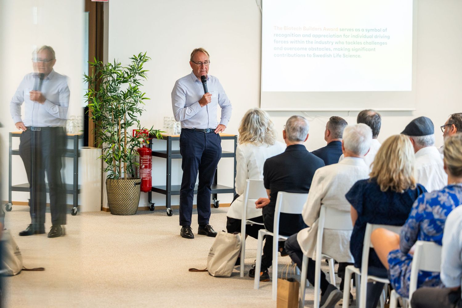 Business speaker presents to seated audience in bright modern conference room setting.