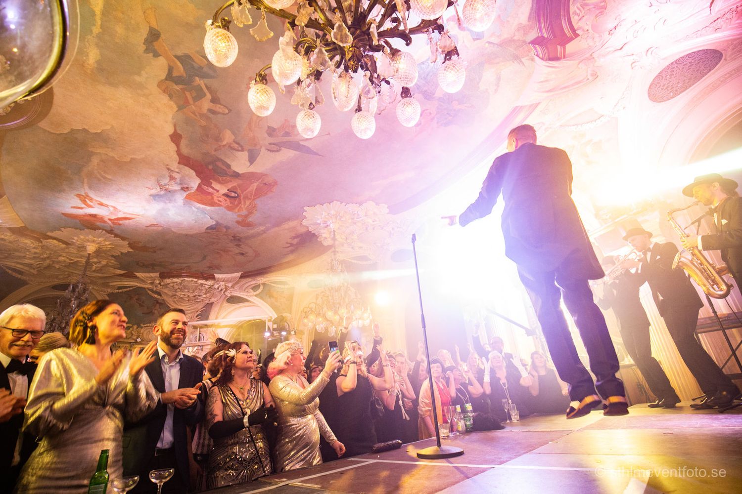Performer on stage entertaining applauding crowd in ornate ballroom with chandelier lighting.
