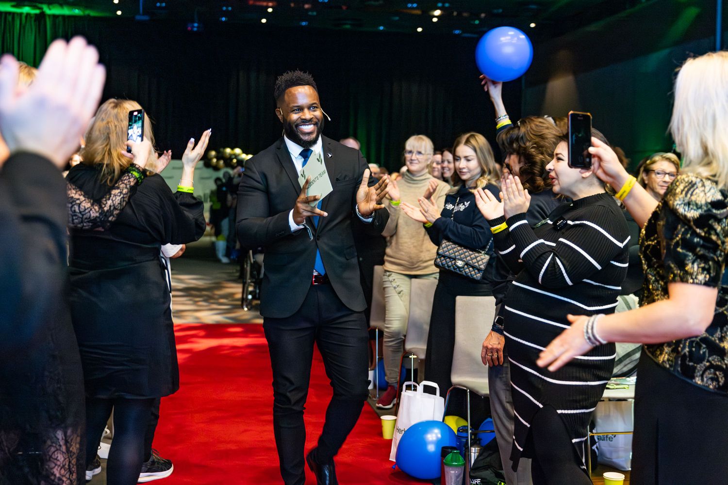 Smiling man in tuxedo walking red carpet surrounded by applauding guests at gala event.