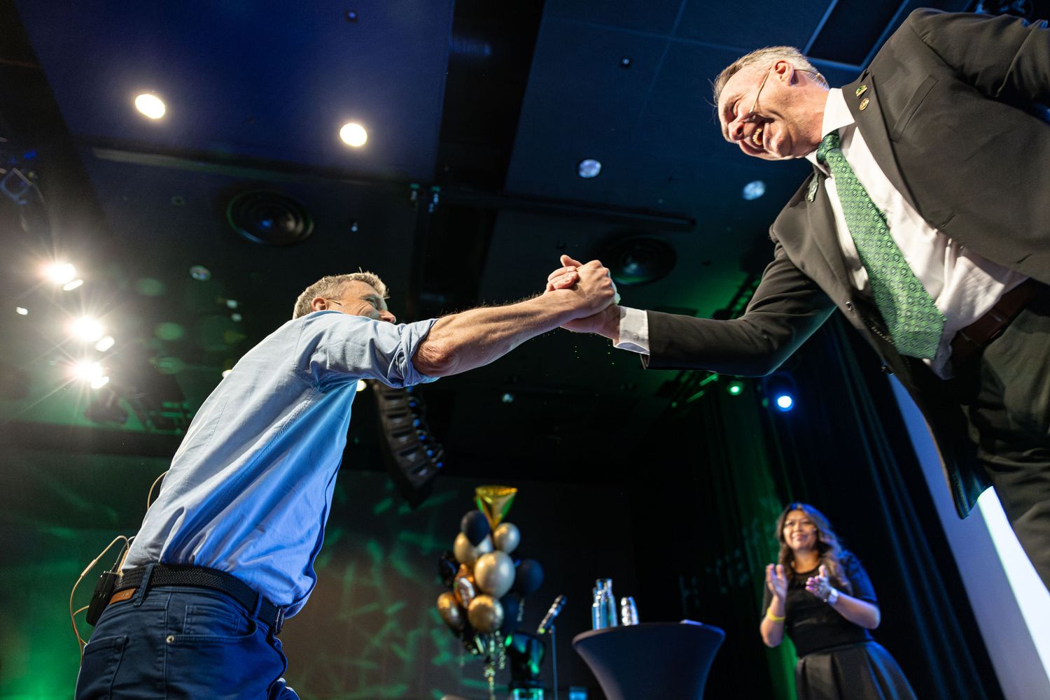Two men share an enthusiastic handshake on a colorful event stage with balloons in the background.