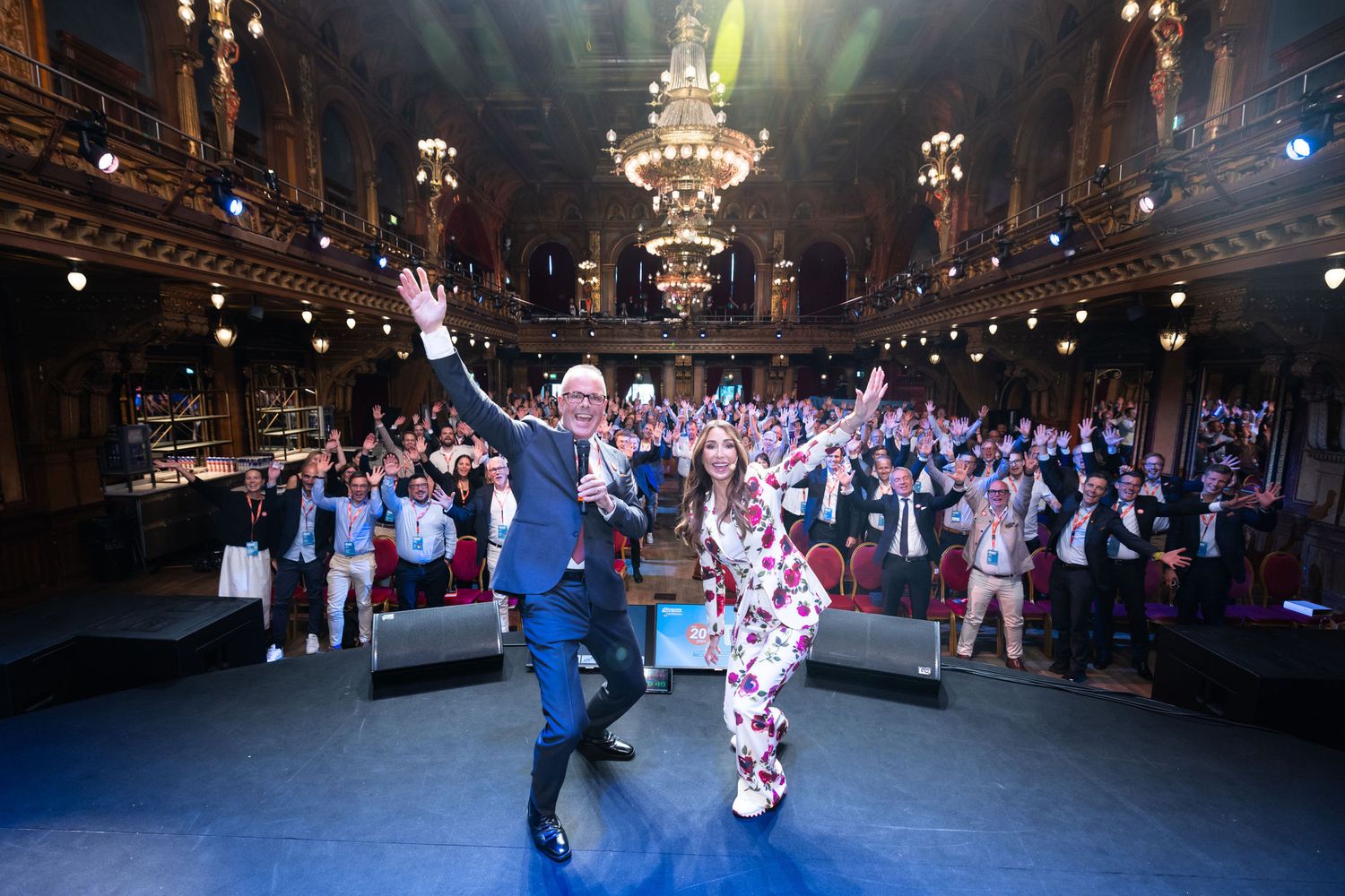 Two enthusiastic presenters wave to a cheering crowd in an ornate grand ballroom at a gala event.