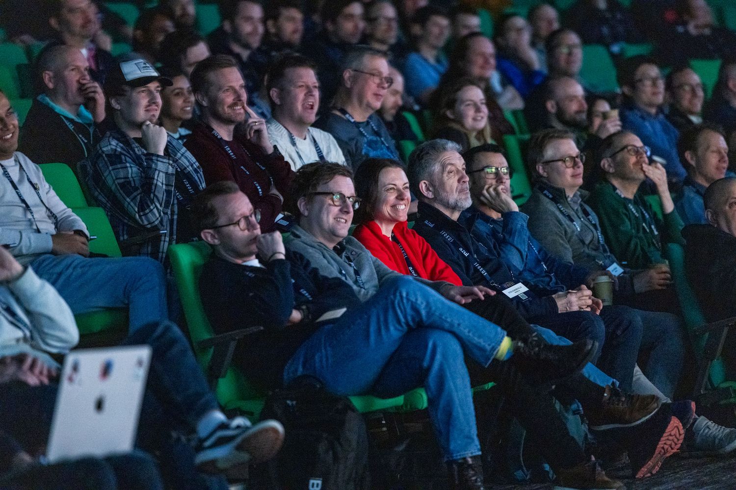 Engaged audience members smile and laugh while watching a presentation in a green-seated auditorium.