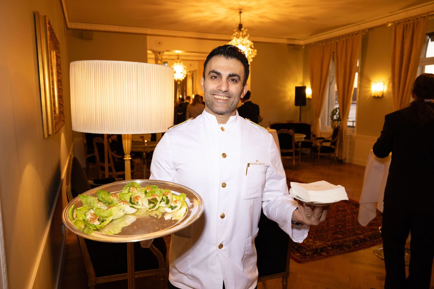 Smiling chef in white uniform proudly presents an elegant green salad dish on a decorative plate.