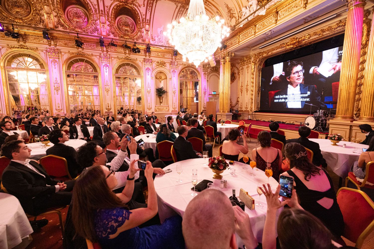 Guests attend a formal awards gala dinner in an ornate ballroom with crystal chandeliers.