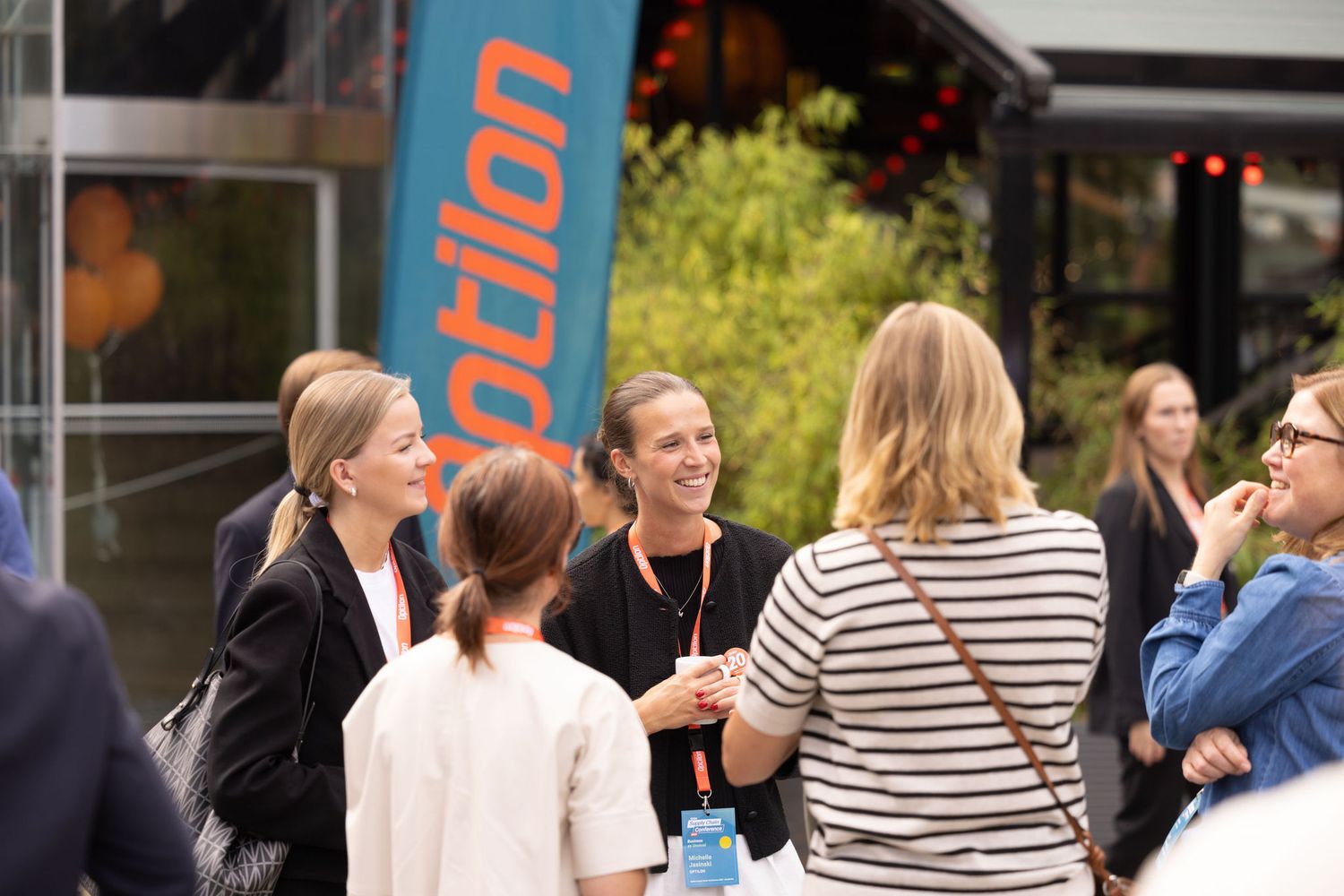 Professional women network and chat outdoors near an Optilon branded banner at a corporate event.