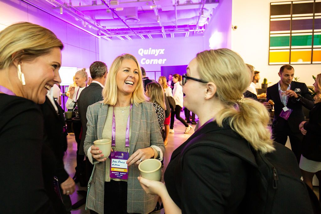 Three women laugh and chat at a corporate networking event with purple lighting and conference lanyards.