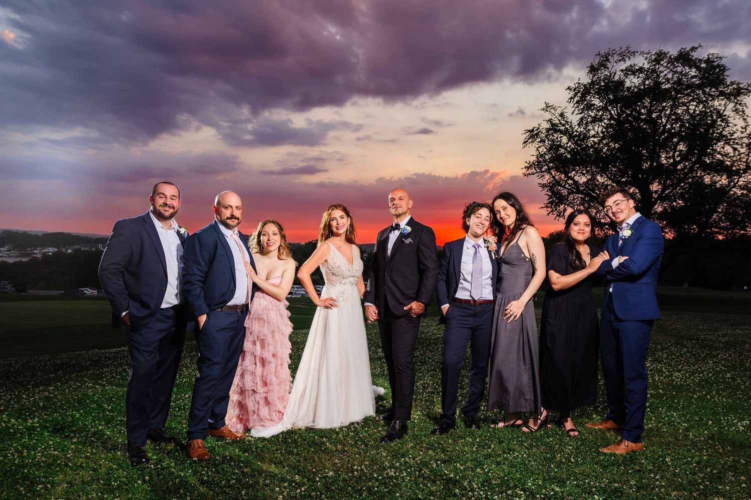 Bride and groom with wedding party standing on a hilltop lawn at sunset with a dramatic purple and orange sky.