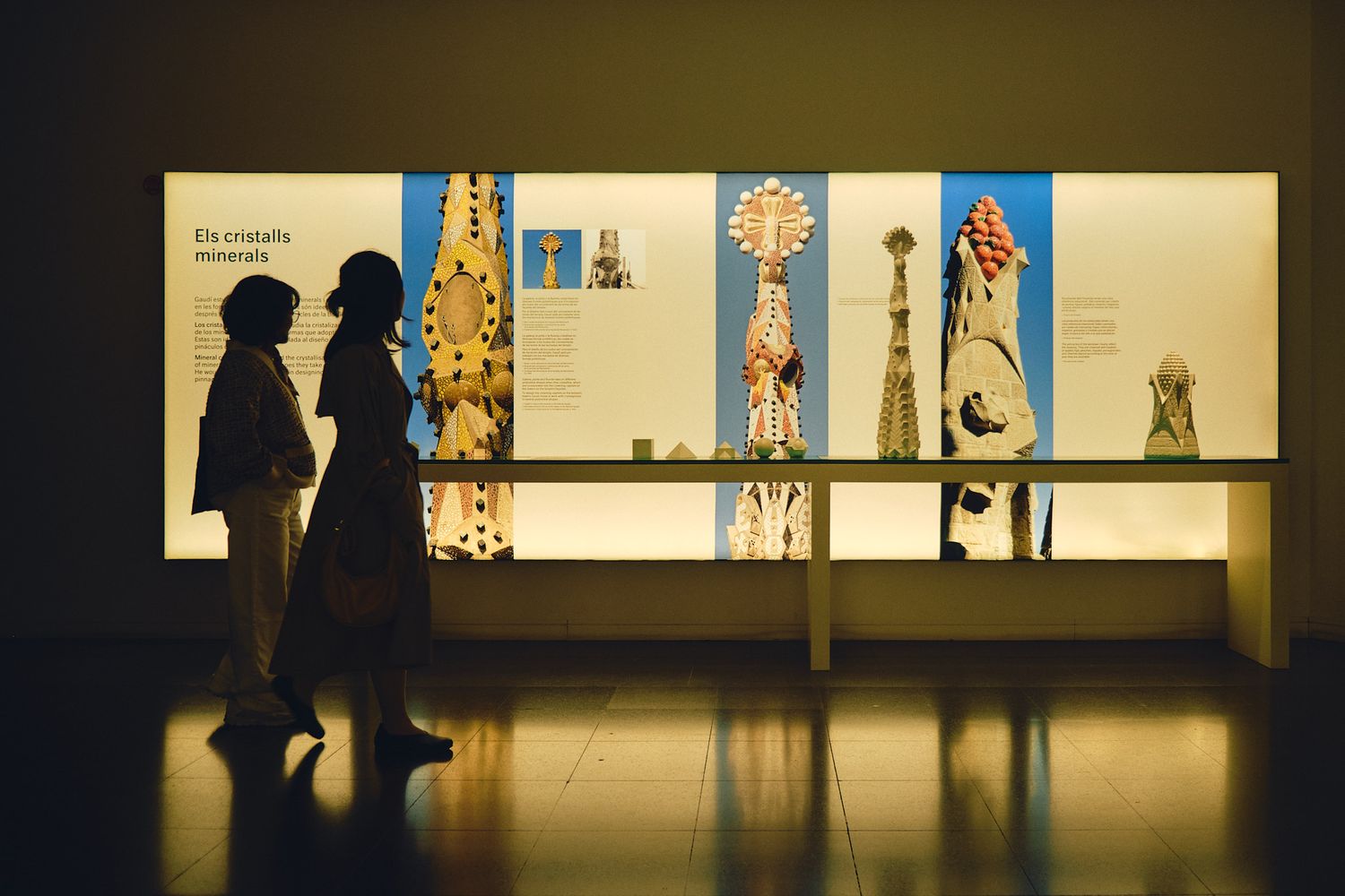 Two silhouetted visitors admire illuminated architectural models and sculptures in a dimly lit museum exhibition gallery.