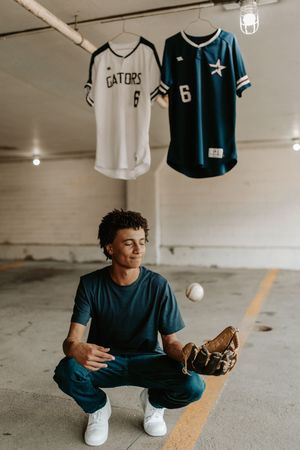 Baseball player crouches in parking garage with Gators #6 jerseys hanging behind him.