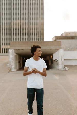 Young man in white t-shirt and jeans smiling outdoors near urban concrete buildings on a city plaza.