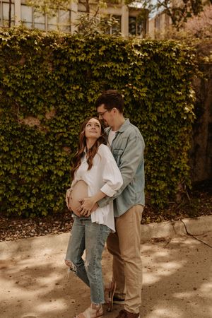 Pregnant couple embracing outdoors against ivy-covered wall, woman in white top and jeans, man in green shirt and khakis.