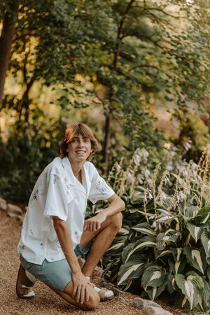 Smiling teen boy in white floral shirt and blue shorts kneeling beside garden plants on a sunny day.