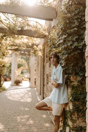 Young person in light blue shirt and white shorts leaning against ivy-covered stone wall in sunlit garden pergola.