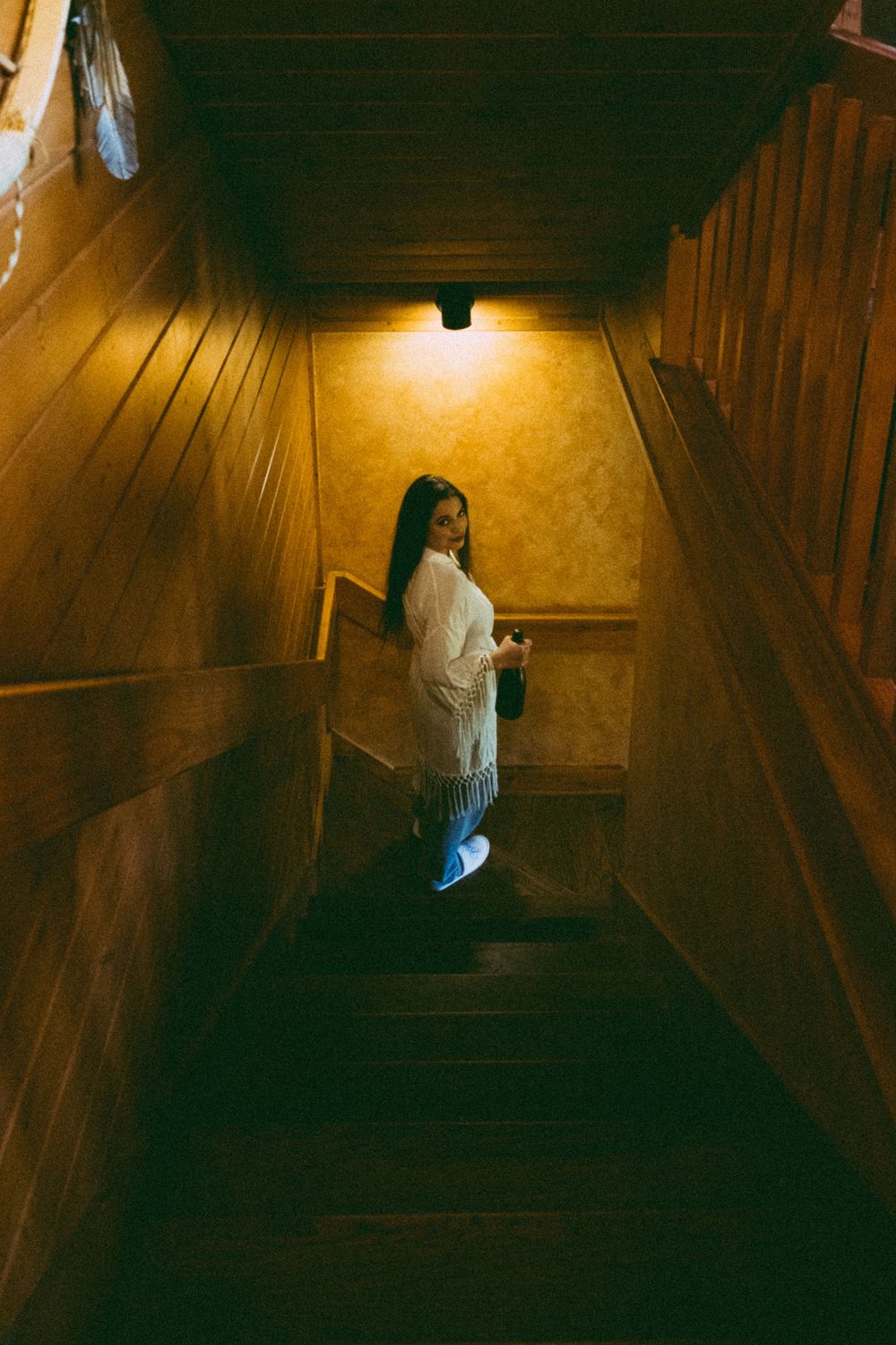 Woman in white fringe top holding bottle on wooden staircase lit by warm overhead light.
