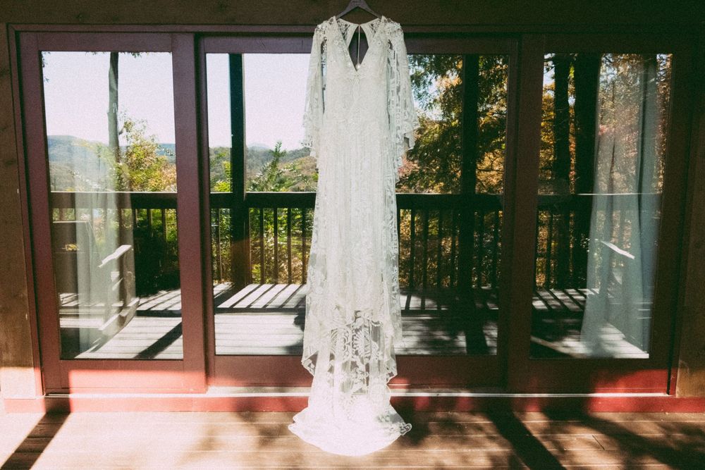 White lace wedding dress hanging on wooden French doors with scenic mountain and forest view in background.