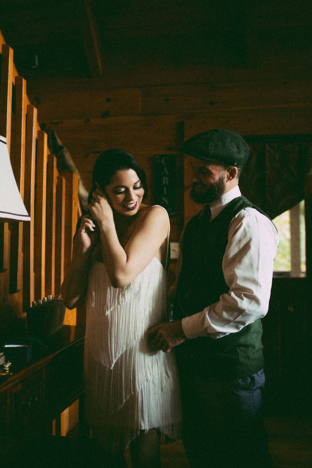 A couple shares a joyful moment inside a rustic cabin, bride in fringe dress and groom in vest and flat cap.