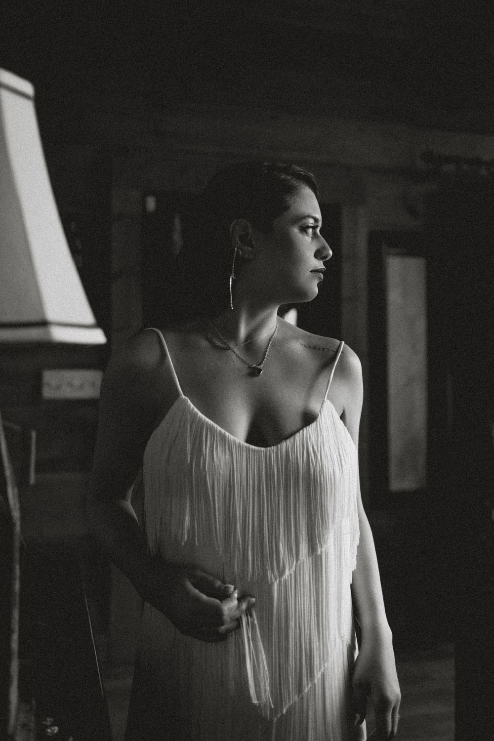 Black and white photo of a woman in a white fringe dress with hoop earrings in a dimly lit room.