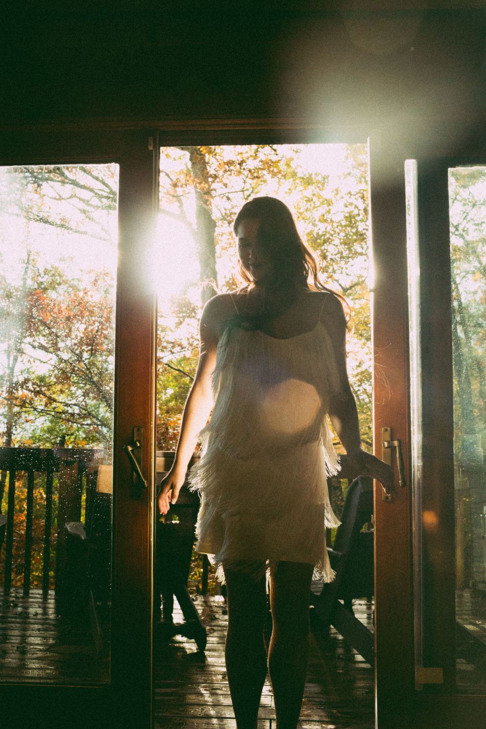 Silhouetted woman in white dress standing in sunlit doorway surrounded by autumn trees.