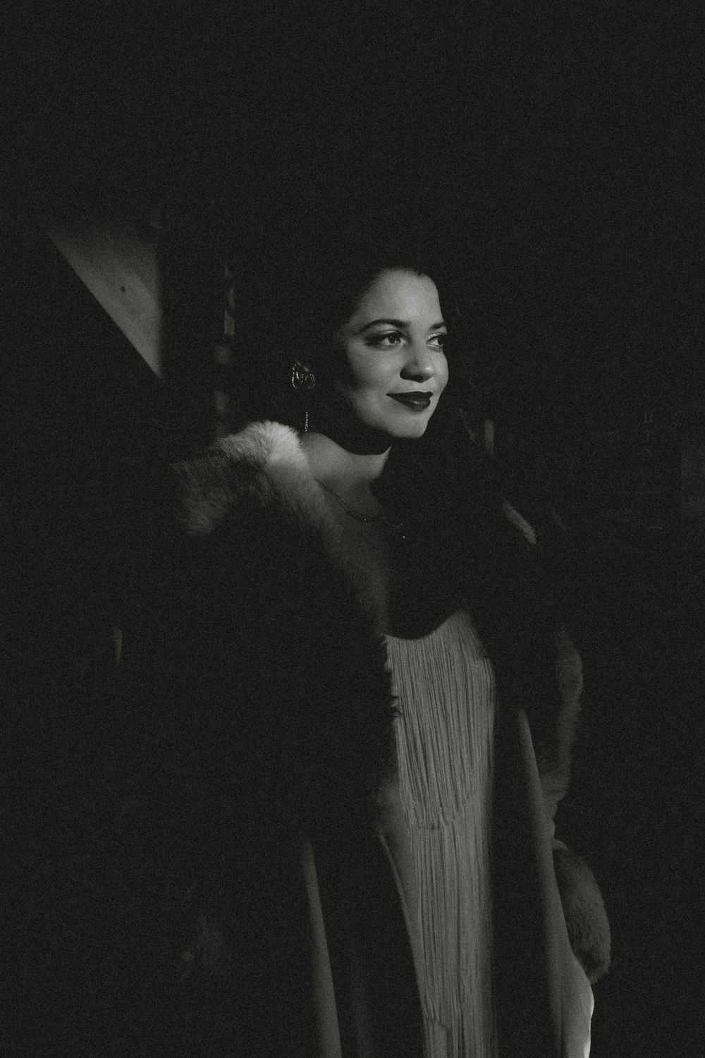 Black and white portrait of a woman in a fur coat and hoop earrings, smiling softly in dim lighting.