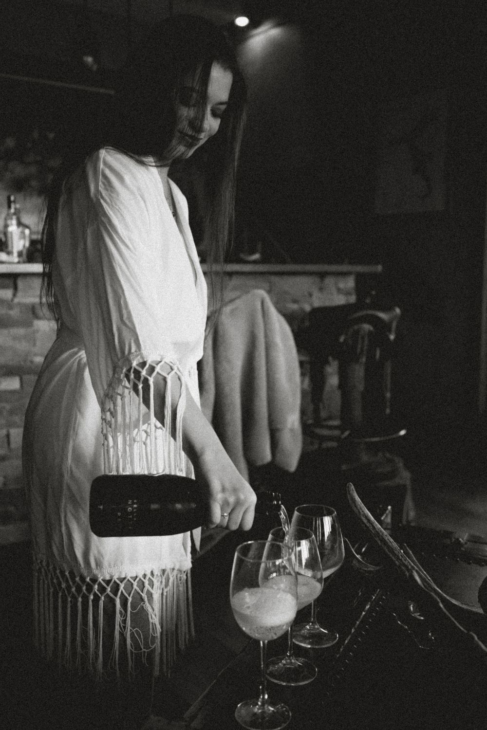 Woman in white fringed dress pouring champagne into wine glasses at a bar in black and white photo.