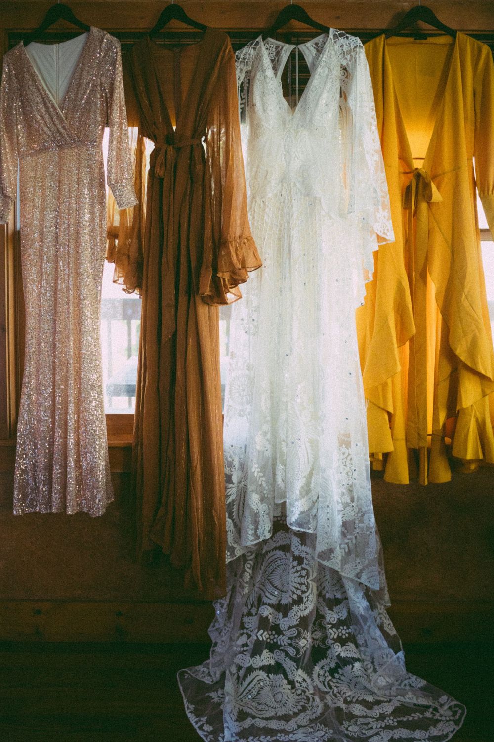 White lace wedding gown hangs center among gold sequin and yellow bridesmaid dresses on wooden hangers indoors.